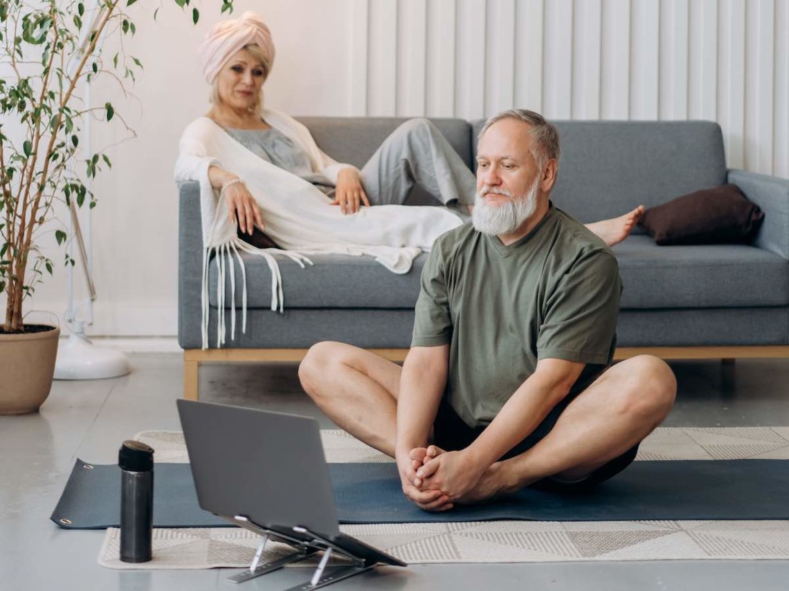 man sitting cross legged - doing yoga with computer on floor with wife on couch behind
